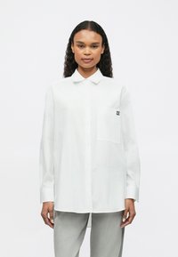 Cămașă albă cu guler ascuțit, un singur buzunar pe piept și logo de text negru pe buzunar. Mâneci lungi și o croială lejeră.