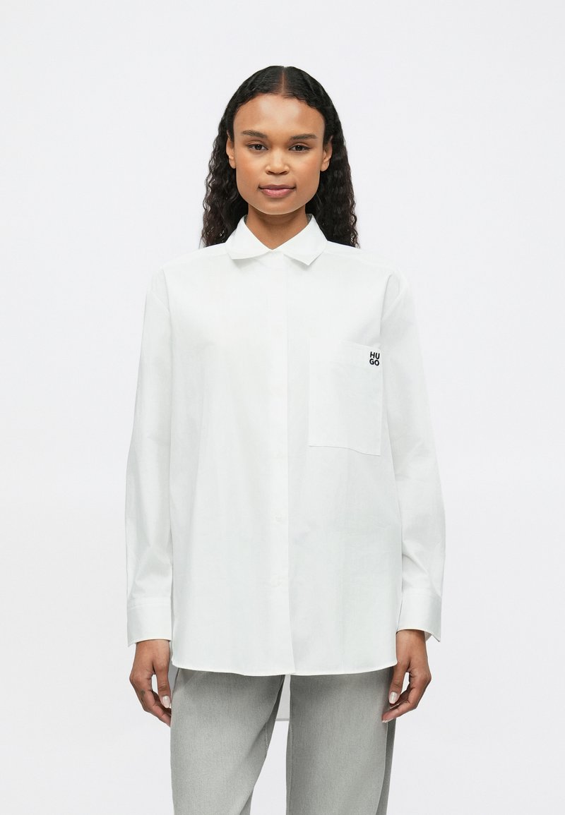 Cămașă albă cu guler ascuțit, un singur buzunar pe piept și logo de text negru pe buzunar. Mâneci lungi și o croială lejeră.