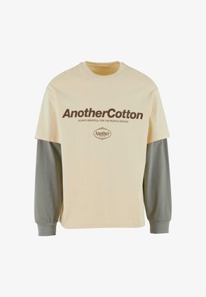 Bežna majica s kratkimi rokavi in sivo dolgimi rokavi. Ponaša se z drznim rjavim napisom: "AnotherCotton" in "VEDNO HVALEŽEN ZA LJUDE OKOLI".