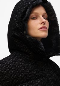 Zwarte gewatteerde jas met een faux fur gevoerde capuchon, uitgerust met een textuurpatroon, knoopsluiting en een soepele stof met verhoogde letters.