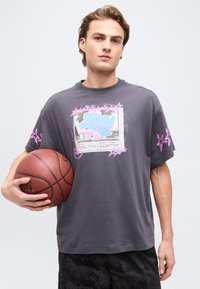 Νεαρός κρατάει ένα μπάσκετ και φοράει γκρι μπλουζάκι με ροζ αστέρια και κείμενο, στέκεται μπροστά σε ένα απλό φωτεινό φόντο.