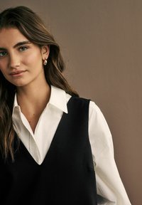 Gilet noir sur une chemise blanche à col, avec un design en V, texture lisse et tissu doux. Les cheveux tombent librement sur les épaules.