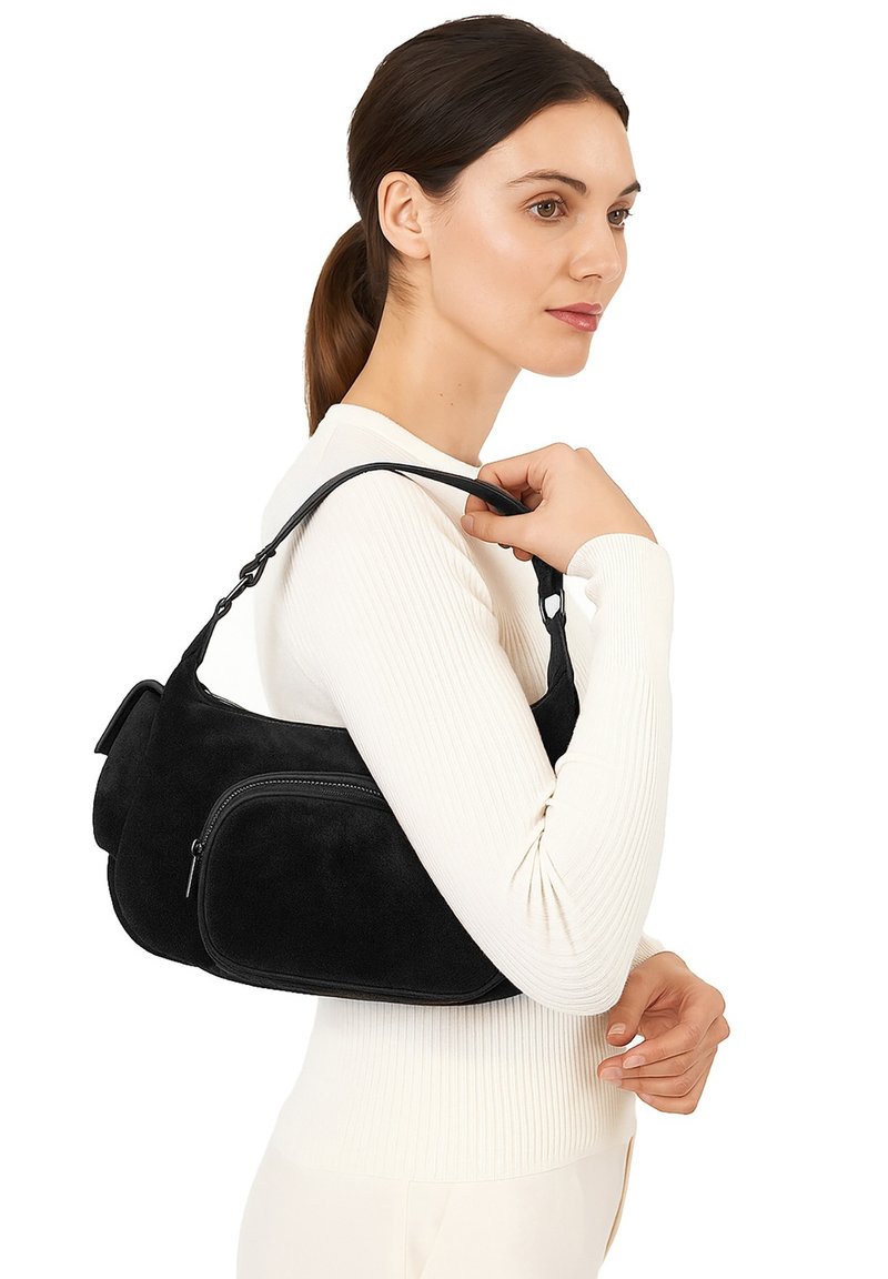 Borsa a tracolla in pelle scamosciata nera con una forma strutturata, dotata di una tasca frontale con zip e spallacci in pelle liscia. Indossata sopra un outfit bianco.