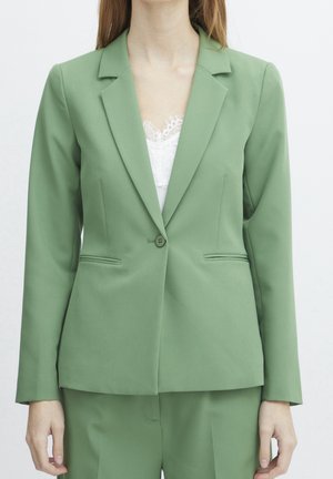 Kvinde iført en lysegrøn skræddersyet jakke over en hvid top med blonder, med matchende lysegrønne bukser synlige nedenunder.