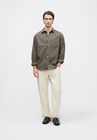 Conjunto informal que incluye una camisa de botones en oliva oscuro con un bolsillo frontal, combinada con pantalones de pierna ancha en beige claro y zapatos negros de slip-on.