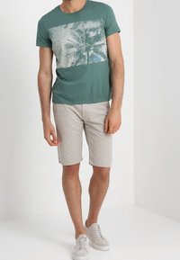 Vihreä puuvillapaita, jossa on grafiikka palmuista, yhdistettynä vaaleanbeigeihin shortseihin ja valkoisiin lenkkareihin, seisoo neutraalin taustan edessä.