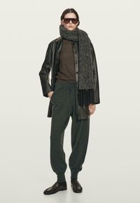 Vrouw in een oversized donkerbruine leren jack, grijze wollen sjaal, bruine top, grijze sweatpants, zwarte loafers en grote zonnebril.