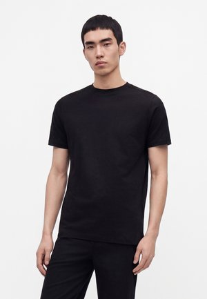 Jeune homme aux cheveux courts et noirs portant un t-shirt noir uni à manches courtes et un pantalon noir sur fond blanc.