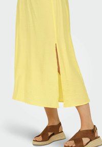Vestido amarillo de longitud a la rodilla con una abertura lateral; presenta una textura suave. Sandalias de cuña marrones con diseño de tiras cruzadas y hebilla en el tobillo.