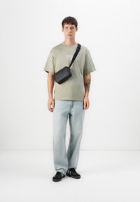 Ung mand iført olivengrønt t-shirt, lyseblå jeans, sorte sneakers og sort crossover taske, stående foran en hvid baggrund.