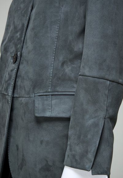 Manga y bolsillo de chaqueta de ante gris oscuro con costuras visibles y un botón negro sobre tela lisa.