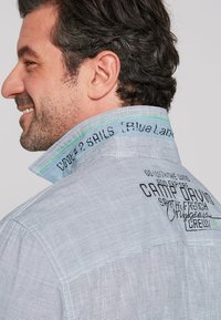 Camisa de botones a rayas con un diseño azul y blanco; cuenta con texto bordado en el cuello y en la parte trasera. Tela de algodón con una textura suave.