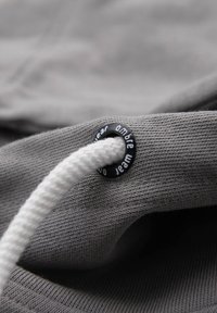 Gros plan sur un tissu gris avec un cordon tressé blanc passant à travers un œillet en métal noir portant l'inscription "ambre wear".