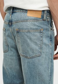 Οπίσθια όψη ενός ατόμου που φοράει τζιν σε ανοιχτό γαλάζιο χρώμα, με ορατές πίσω τσέπες και ταμπελάκι από καφέ δέρμα με το λογότυπο στη ζώνη της μέσης.