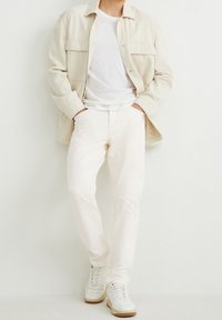 Camisa de botones crema sobre una camiseta blanca, combinada con pantalones beige claros y zapatillas blancas con suelas marrones. Diseño simple y casual.