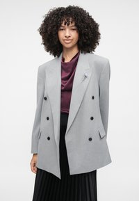 Harmaa, kaksirivinen bleiseri mustilla napeilla, terävillä läpäillä ja teksturoidulla kankaalla, yhdistettynä purppuranväriseen satiinitoppaukseen ja laskostettuun mustaan hameeseen.