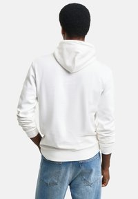 Sudadera blanca hecha de tejido suave con capucha ajustable, puños acanalados y dobladillo. Combinada con jeans de mezclilla azules.