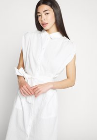 Vestido branco de camisa com mangas curtas e estruturadas, frente com botões, colarinho, cinto atado e bolsos frontais, apresentando uma textura de riscas verticais subtil.