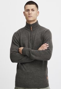 Pull à col zippé gris foncé avec des poignets et un ourlet côtelés, fabriqué en tissu tricoté texturé, avec un patch en cuir au bas.