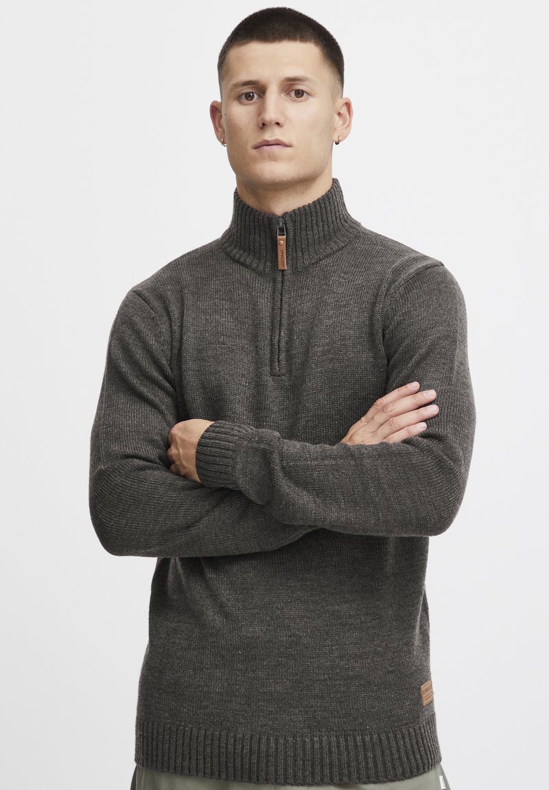 Pull à col zippé gris foncé avec des poignets et un ourlet côtelés, fabriqué en tissu tricoté texturé, avec un patch en cuir au bas.