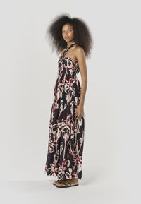 Vestido maxi negro con estampado floral y abstracto en rojo y crema. Diseño con cuello halter, tela fluida y dobladillo en capas. Sandalias planas con tiras.