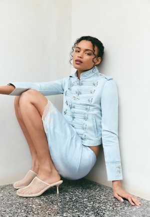 Ung kvinde med krøllet hår sidder op ad en hvid væg iført lyseblå jakke og nederdel samt beige net-højhælede mules på terrazzo-gulv.