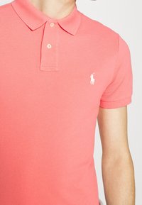 Korallfärgad polo skjorta i bomull, med klassisk krage, två knappar och en liten vit logotyp på vänster bröst. Strukturerad tyg.