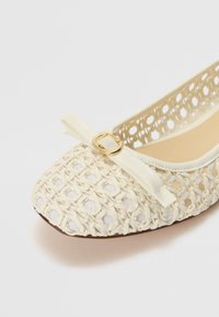 Zapato plano de color crema con un diseño de tejido entrelazado. Presenta un pequeño lazo decorativo y un detalle de hardware dorado en la parte delantera.