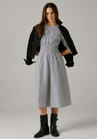 Mladi žena u midi haljini s prugama i stegnutim strukom, s crnom jaknom prebačenom preko ramena i crnim gležnjačama, stoji na neutralnoj pozadini.