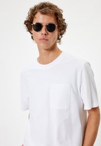 Hvid t-shirt med rund hals og korte ærmer samt en brystlomme, parret med runde sorte solbriller. Glat tekstur og afslappet design.