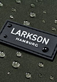 Grön texturerad tyg med vattendroppar, med en svart rektangulär etikett med texten "LARKSON HAMBURG" i vita bokstäver.