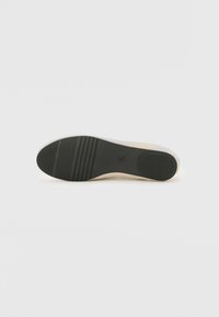 Die Schuhsohle verfügt über eine glatte schwarze Gummiunterseite mit horizontalen Rillen für den Grip, umgeben von einem hellbeigen Rand und einem strukturierten Design.