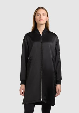 Frau trägt lange schwarze Bomberjacke mit Frontreißverschluss und Seitentaschen, steht vor einem einfachen hellgrauen Hintergrund.