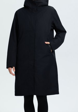 Person iført en lang sort hættejakke med hænder delvist i sidelommer mod en ensartet lys baggrund.