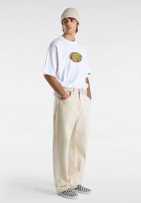 Witte t-shirt met korte mouwen en een grafisch logo, beige wijde broek en zwart-witte geruite instappers. Gebreide beige beanie.