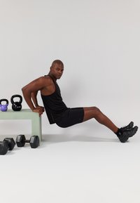 T-shirt preta e calções usados por uma pessoa a realizar mergulhos de tríceps num banco verde, com halteres e kettlebells nas proximidades.