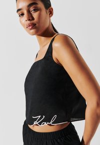 Crni bezrukavni crop top s kvadratnim izrezom i bijelim vezom s potpisom "Karl" na rubu. Glatka tekstura i opušteni kroj.