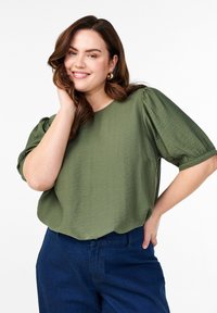 Top olive vert à manches courtes avec une finition texturée, coupe ample, encolure ronde et manches froncées. Associé à un pantalon bleu foncé.