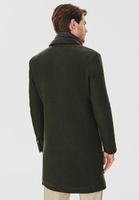 Ciemnozielony wełniany płaszcz o gładkim, teksturowanym wykończeniu. Posiada klasyczny kołnierz i zapięcia na guziki na mankietach, zaprezentowany z tyłu.