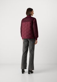 Giacca cucita color borgogna con colletto alto e polsini elasticizzati, abbinata a jeans a gamba larga grigio scuro. Tessuto morbido, dettaglio di cuciture geometriche.