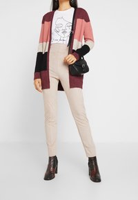 Stickad cardigan i vinrött, rosa och svarta ränder, lager över en vit grafisk t-shirt. Kombinerad med beige byxor och svarta klackstövlar.