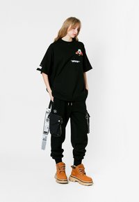 Camiseta negra de gran tamaño con estampado floral y texto, combinada con joggers negros con bolsillos laterales y una correa negra. Botas marrones con suelas gruesas.