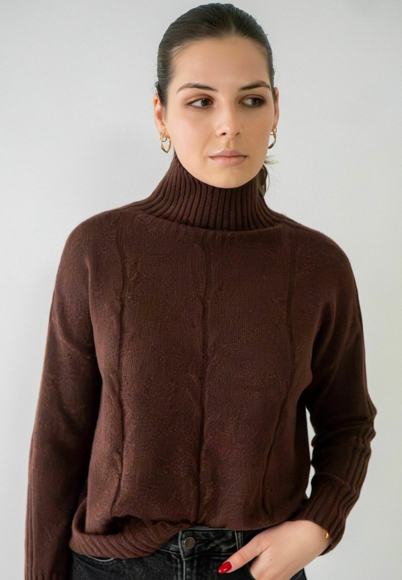 Pull à col roulé marron avec un motif de tricot en cable texturé, des poignets côtelés et une coupe décontractée, associé à un jean en denim foncé.