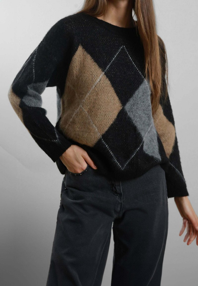 Donna che indossa un maglione con motivo argyle nero, beige e grigio, con pantaloni neri, una mano in tasca, su uno sfondo grigio uniforme.