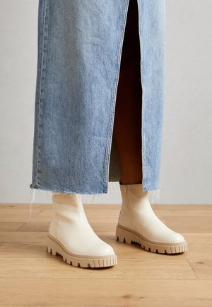 Beige ankle boots with a textured platform sole and side zipper, made of smooth material, paired with a frayed denim maxi skirt.