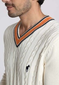 Pull en maille blanche avec un décolleté en V profond. Présente des bordures à rayures orange et noires, orné d'un petit logo de cheval noir.