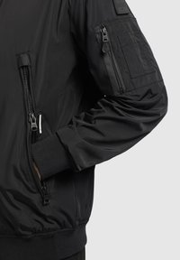 Schwarze Bomberjacke aus einem glatten Außenmaterial, mit gerippten Bündchen, Reißverschlusstaschen und einem Aufnäher am Ärmel, die ein schlankes Design aufweist.