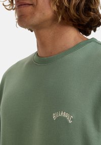 Grüner Sweatshirt mit Rundhalsausschnitt, strukturiertem Stoff und einem beigefarbenen gestickten Logo "BILLABONG" auf der Brust.