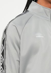 Acercamiento a una persona que lleva puesta una chaqueta Umbro de color gris claro con rayas de patrones de diamante en blanco y negro en el hombro.