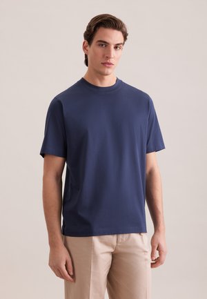 Junger Mann mit dunklen Haaren, der ein schlichtes dunkelblaues T-Shirt mit kurzen Ärmeln und beige Hosen trägt, steht vor einem hellen, neutralen Hintergrund.